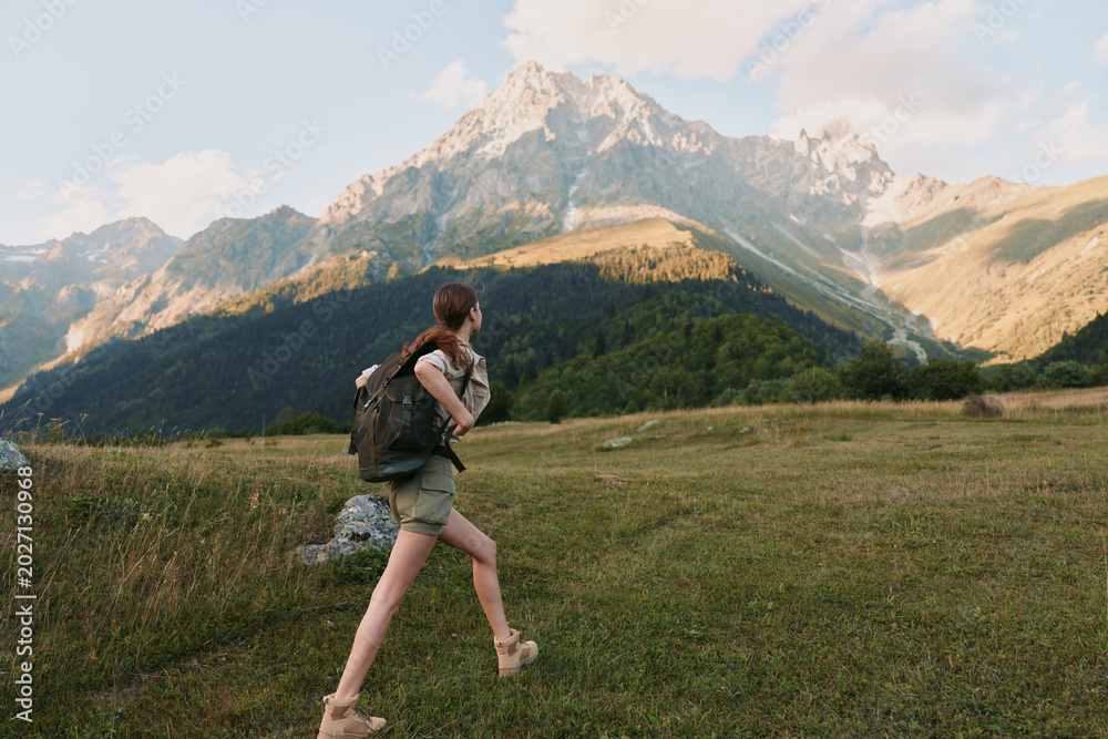 Fototapeta premium A woman with a backpack hikes through a grassy meadow towards tall mountains under a partly cloudy sky. The scene captures outdoor adventure and lifestyle in nature.