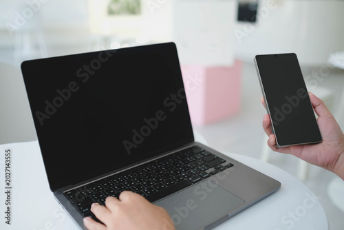 An over the shoulder view of a person using a laptop and holding a smartphone that both feature blank black mockup screens for custom content insertion.