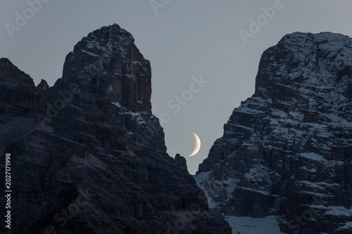 Crescent moon setting between snow-covered mountain peaks during twilight in late autumn. Monte Cristallo, South Tyrol, Italy.