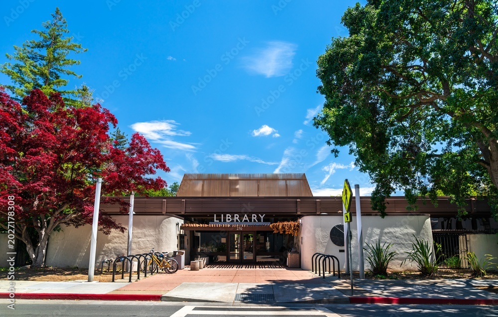 Fototapeta premium Palo Alto, California - May 29, 2023: Exterior view of the Palo Alto City Library Downtown Branch building, featuring bright red and green foliage against a blue sky in Silicon Valley, United States