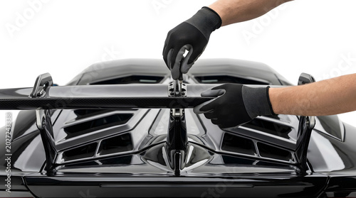 Gloved hands assembling the central strut of a carbon fiber rear wing on a black luxury supercar