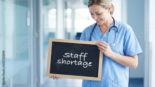 Smiling healthcare worker in scrubs holding a chalkboard with staff shortage text, nurse staff smiling with a message sign in hospital corridor, workforce healthcare medical issue concept