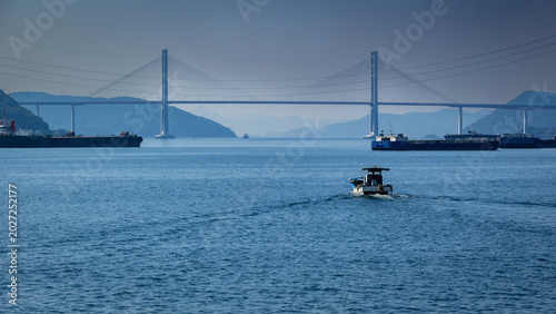 Obraz na płótnie Cable-stayed bridge over calm blue sea with cargo ships and small fishing boat,