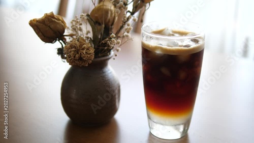 Cinematic close-up of iced Americano black coffee with rich crema foam layer next to a dried flower vase on a rustic wooden table
