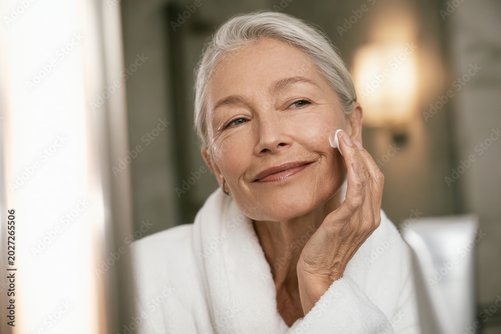 Fototapeta premium Mature woman applying sunscreen on face in bathroom mirror
