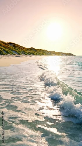 Golden sand beach with lush green dunes dotted with tiny wildflowers and small pebbles. Gentle ocean waves lap the shore under a clear pastel sky as the sun sets, casting a warm glow on the landscape.