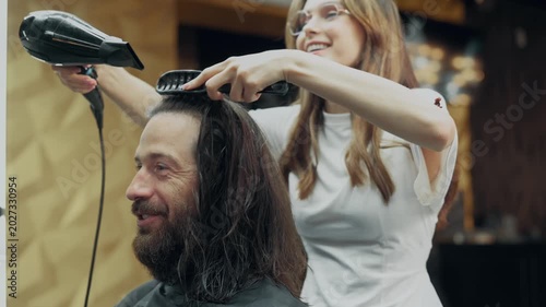 Female barber drying hair of male client with hairdryer during professional hairstyling procedure in modern barbershop interior