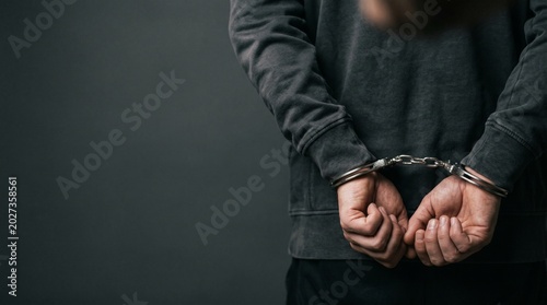 Arrested suspect in handcuffs behind his back standing against a dark grey background with copy space