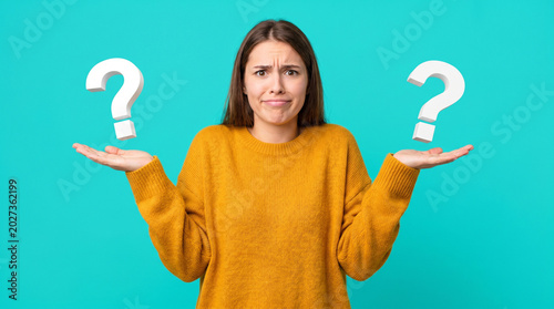 Young woman looking confused while holding question marks on blue background  