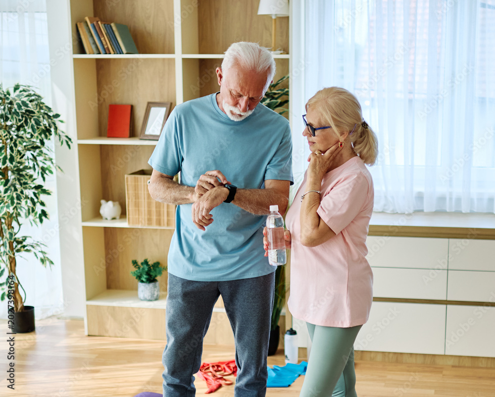 Naklejka premium Smiling active senior couple exercising taking a break on the mat at home or in the gym, senior athlete checking his heart rate and performance with smartwatch