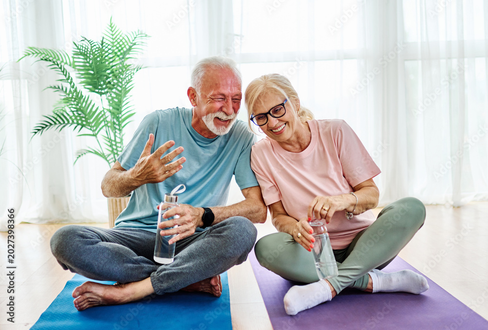 Fototapeta premium Portrait of a senior couple holding water bottle and exercise mat smiling and talking at home healthy living