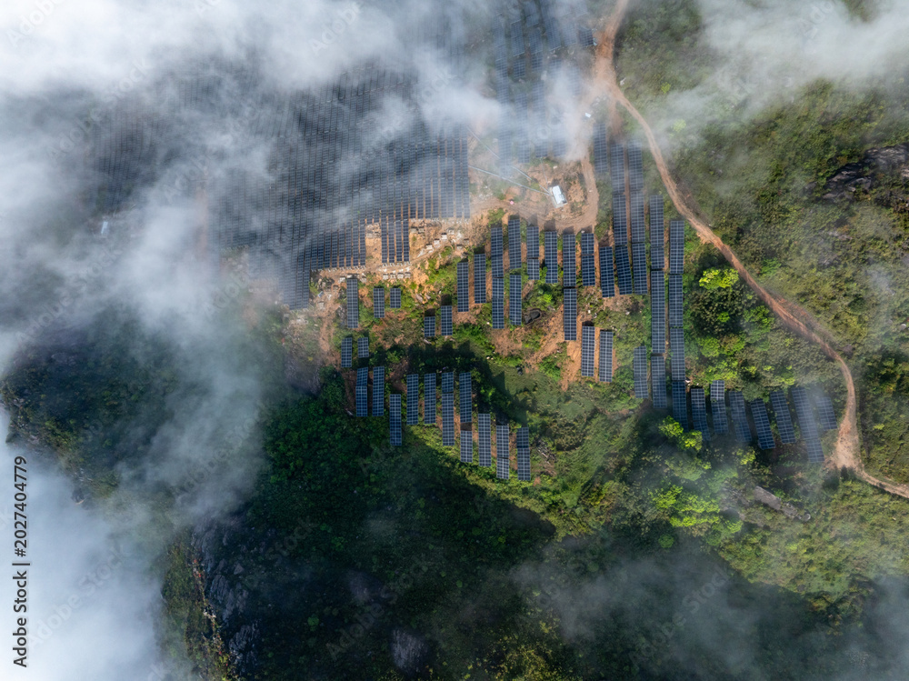 Fototapeta premium Aerial view of solar power station on misty mountain surrounded by clouds and greenery