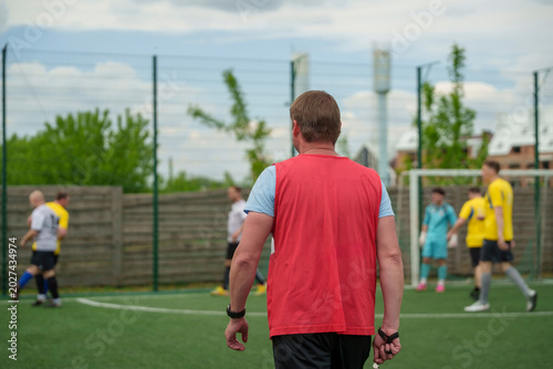 Photos Man in a red vest walking onto a green soccer field during a sunny daytime match, amateur football referee coach, sports activity