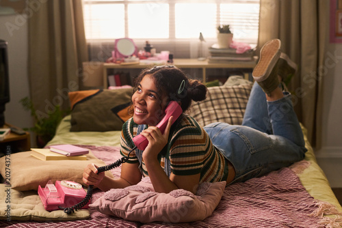 Teenage girl lying on bed and talking on retro landline phone, expressing friendship and nostalgic communication. Ideal for youth lifestyle, teen culture, social connection marketing
