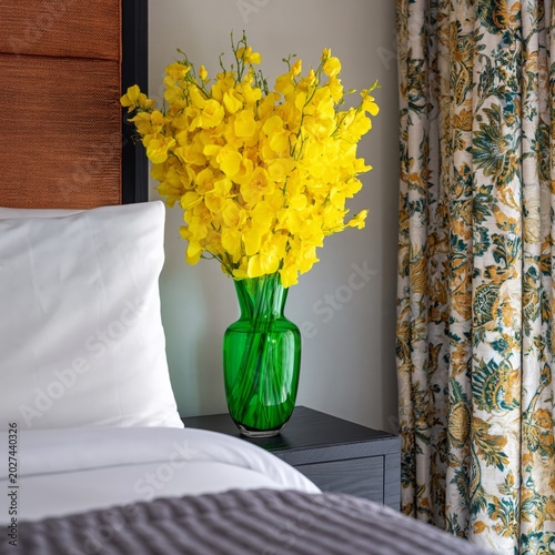 Bright Yellow Flowers in Green Vase on Nightstand Next to Bed in Room With Pa...
