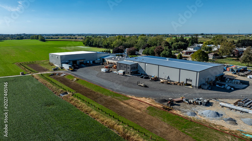 Large farm buildings are seen in a rural setting. Green fields surround the area, and equipment is parked nearby. The sky is clear and bright.