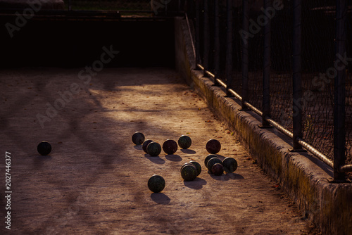 Fotografie A moody, low-angle shot of a traditional Mediterranean bocce court during the golden hour, where scattered bocce balls cast long shadows on the dirt ground
