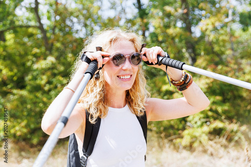 Happy woman hiking while adjusting sunglasses with trekking poles in sunny forest, joyful outdoor travel and active lifestyle concept