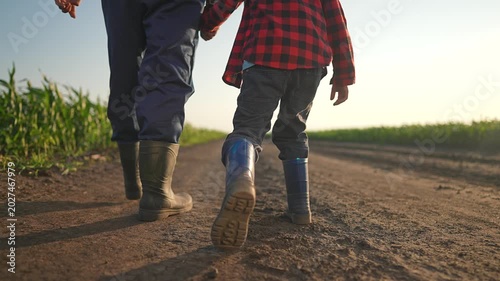 Farmer and boy walking dirt road at sunset. Man leads child with gentle care near summer field. Boots crush warm ground. Walking side by side builds bond. Road stretches far under soft evening sky.
