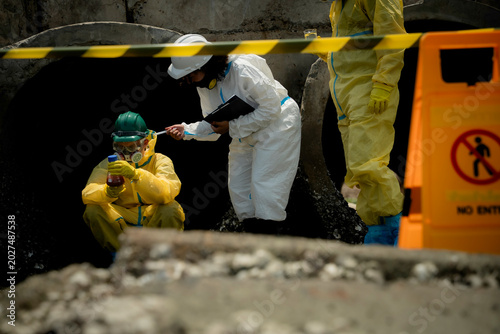 Hazardous wastewater contamination and restricted area concept. Environmental inspectors in hazmat suits monitoring polluted industrial drainage site with no entry warning sign
