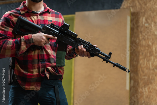 Foto Man in plaid shirt holding tactical rifle with collimator sight at indoor shooti