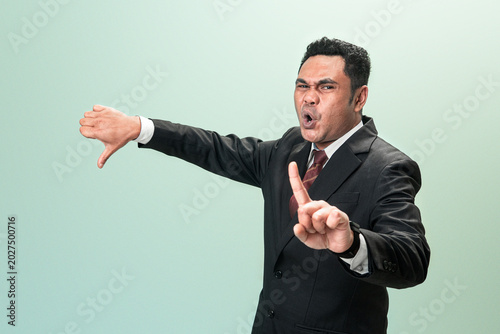 Foto Disgusted Asian businessman in suit showing thumbs down and stop finger gesture in disapproval against green studio background