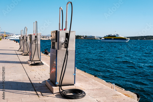 Sleek marine fuel pumps line a sunlit Mediterranean dock. A passenger ferry glides across tranquil azure waters under a clear sky