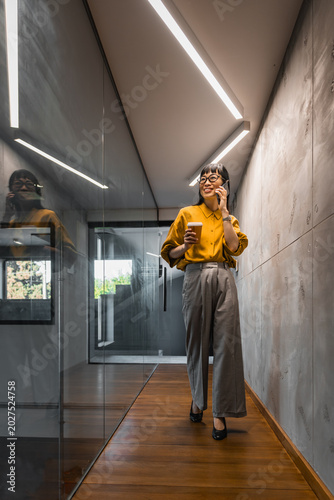 Professional woman walking in modern office hallway talking on phone and holding coffee cup