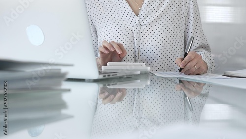 Tableau sur toile Unknown businesswoman using a calculator to crunch numbers and jotting down meticulous notes at a sleek glass desk in a contemporary office, closeup view