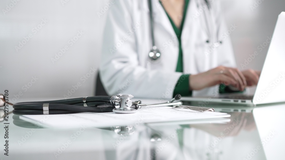 Fototapeta premium Female doctor using technology in healthcare, close up of hands typing on laptop with stethoscope, glasses, and clipboard on desk