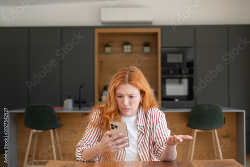 Fotografía Frustrated redhead woman looking at smartphone in modern kitchen, confused by a