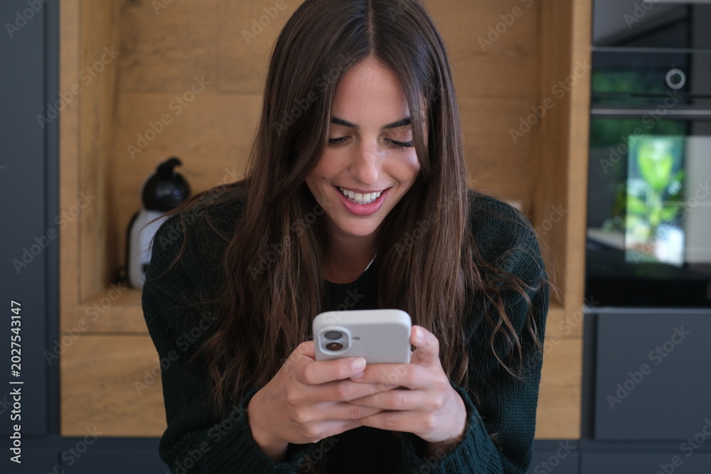Fototapeta premium Young woman smiling and texting on smartphone while relaxing in modern kitchen at home