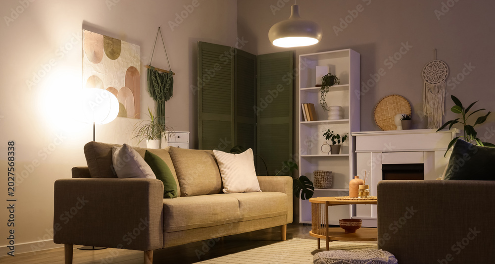 Fototapeta premium Interior of stylish living room with grey sofas, coffee table, fireplace and glowing lamps at evening