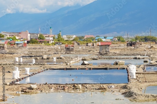 Salt pans stretch across a sunlit, dry landscape. White sacks stand in shallow water, resting on wooden racks. Distant buildings dot the horizon, blending with the terrain