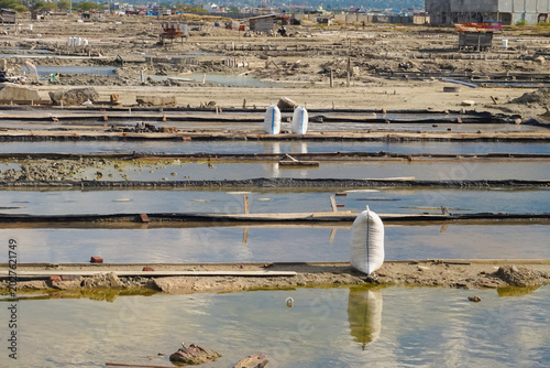 Salt pans stretch across a sunlit, dry landscape. White sacks stand in shallow water, resting on wooden racks. Distant buildings dot the horizon, blending with the terrain