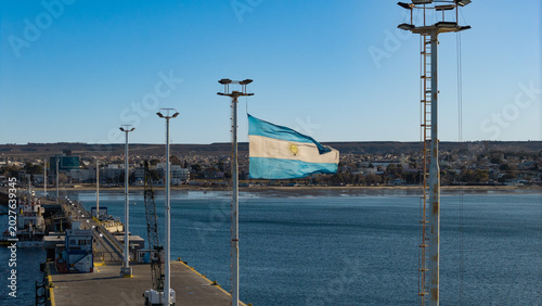 Obraz na plátně Argentine flag waving above Puerto Madryn pier with Atlantic Ocean and Patagonian city background