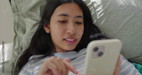 Asian teenage girl lying near on bed, using smartphone, Sunlight filters through sheer curtains in bedroom