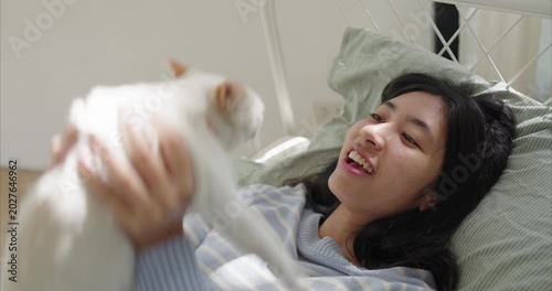 Asian woman lies on a tidy bed ,lifts white cat toward playing, sunlit Warm late afternoon light, Rest, relaxation, good mood concept