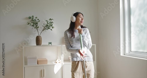 young Asian woman in a striped sweater dances with a mop as a microphone in a bright minimalist home, wearing white headphones