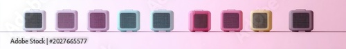 row of nine pastel-colored cube speakers with front grills on a thin shelf against a soft pink background, evoking a playful cheerful mood