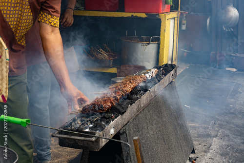 Photos A man is grilling Sate Buntel (culinary specialty from Solo, Central Java, Indonesia), featuring minced goat or lamb meat wrap in fat and grilled over charcoal with billowing smoke
