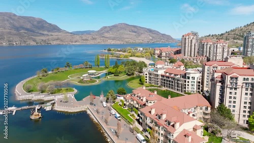 Lakeview of a building complex with coral coloured roofs and man-made landscape at the water's edge, with mountain and water in the background, on a bright and sunny day.
