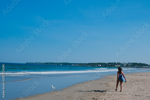 Bild auf Leinwand A woman carrying a bodyboard walks along a sandy ocean beach toward breaking waves under a clear blue sky, with a seagull near the shoreline and a distant coast in view