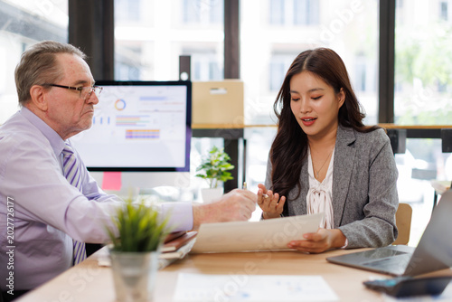 Generations of colleagues collaborate over reports and a laptop displaying charts, mentoring, strategizing finance and planning in a modern corporate office setting
