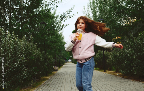 Fotografie Cute smiling girl having fun in the city park, drinking lemonade , street food,  Retro style, nostalgic, dreamy atmosphere, Blurred mootion draws attention to the naturalness of the moment