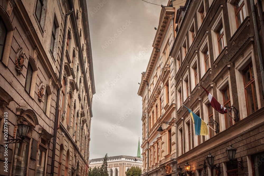 Obraz premium Narrow cobblestone street in Vecriga Vecpilseta, the Old Riga, Latvia, with historic facades, flags, street lamps and a view toward the old town architecture of the Latvian capital.