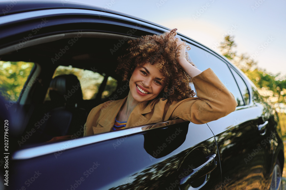 Fototapeta premium A smiling woman leans out of a sleek car window, wearing a casual jacket, as warm sunlight floods the scene. An upbeat lifestyle moment captured outdoors with freedom.