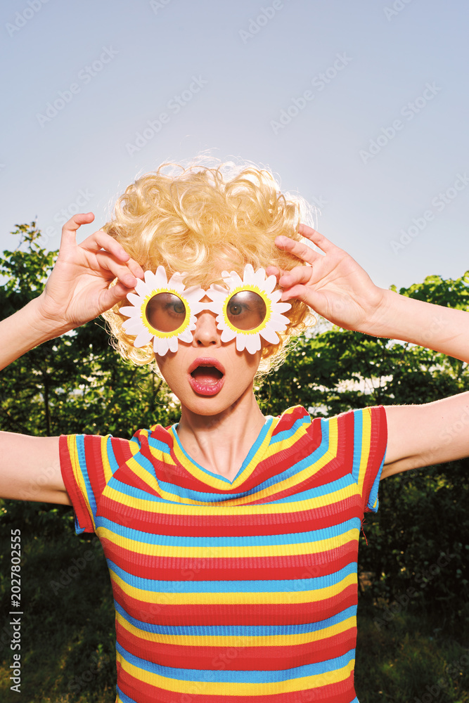 Fototapeta premium Smiling woman with curly blonde hair wearing a striped colorful shirt outdoors on a sunny day, playful retro vibe with flower glasses and vibrant mood.