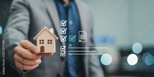 Businessman holding miniature wooden house model with document checklist icon.
