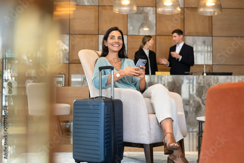 Smiling female hotel guest sitting in reception using smartphone to check messages, confirm reservation, or complete online check-in. Digital services, modern travel app usage, mobile communication